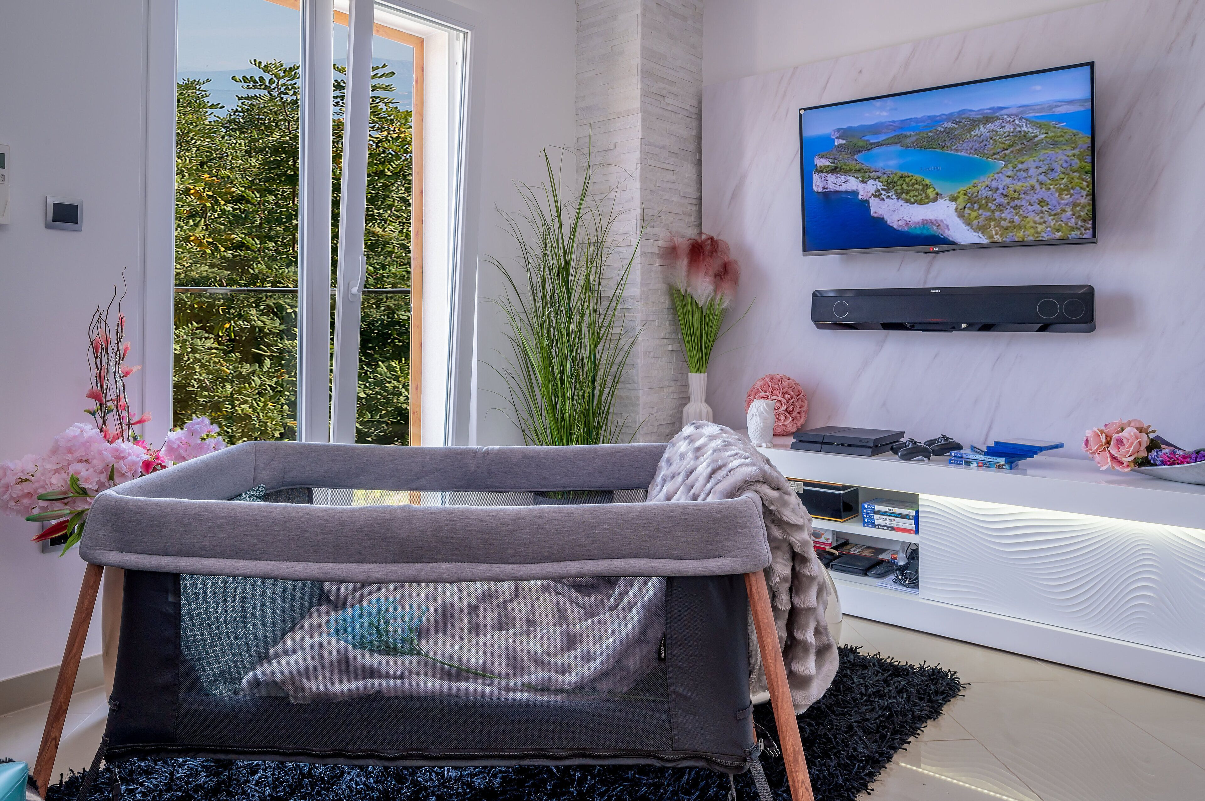 A modern living room with a baby play area, a large window with a view of greenery, a flat-screen TV, and a white entertainment unit.