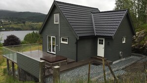 Een donkergroen huis met schuin dak, een balkon en uitzicht op een meer en heuvels.