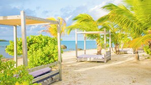 A beach setting with wooden structures, including a canopy and a bed, surrounded by palm trees and a view of the sea.