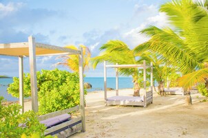 Escena de playa con estructuras de madera, incluyendo una sombrilla y una cama, rodeado de palmeras y vistas al mar.