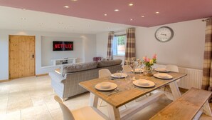 A dining area with a wooden table set for four, a sofa, a TV displaying 'NETFLIX', and a clock on the wall.