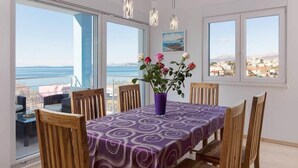 A dining area with a table set for four, large windows offering a view of the sea and buildings, and a vase with flowers on the table.
