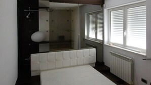 Un dormitorio pequeño y funcional con cama individual, un banco empotrado, un radiador y una ventana con persianas.