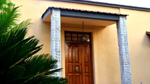 Una puerta de madera con un llamador de latón, flanqueada por dos pilares de piedra, que da paso a un edificio con fachada amarilla y techo de tejas.