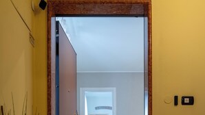 A hotel entrance with a wooden door, a doormat reading "Casa Helena," a potted plant, and a glimpse of a living room with a sofa and a small table.