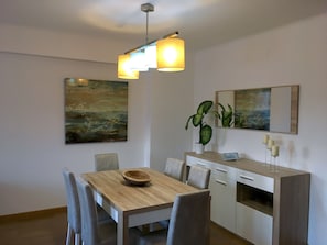 Uma sala de jantar com mesa de madeira, cadeiras, luminária pendente, planta em vaso e obras de arte nas paredes.
