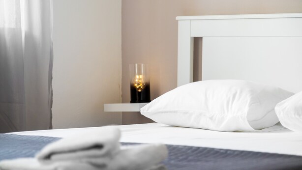 Ein ordentlich gemachtes Bett mit weißer Bettwäsche, ein Nachttisch mit einem Glas Wasser und eine an der Wand befestigte Lampe.
