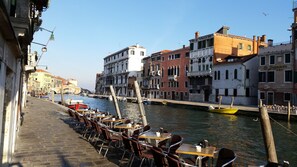 A riverside café with outdoor seating overlooking a canal, historic buildings, and a bridge in the background.