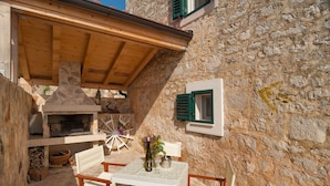 A stone building with a wooden table and chairs, a stone fireplace, and a window with green shutters.