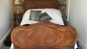 Un dormitorio con pared de piedra, una cama de madera con cabecero curvo, una mesita de noche pequeña con lámpara y suelo de baldosas grises.