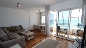 Un salon moderne avec un canapé gris, une table basse et un téléviseur. On y trouve également un coin repas avec une table et des chaises, ainsi qu’un balcon offrant une vue sur l’océan.