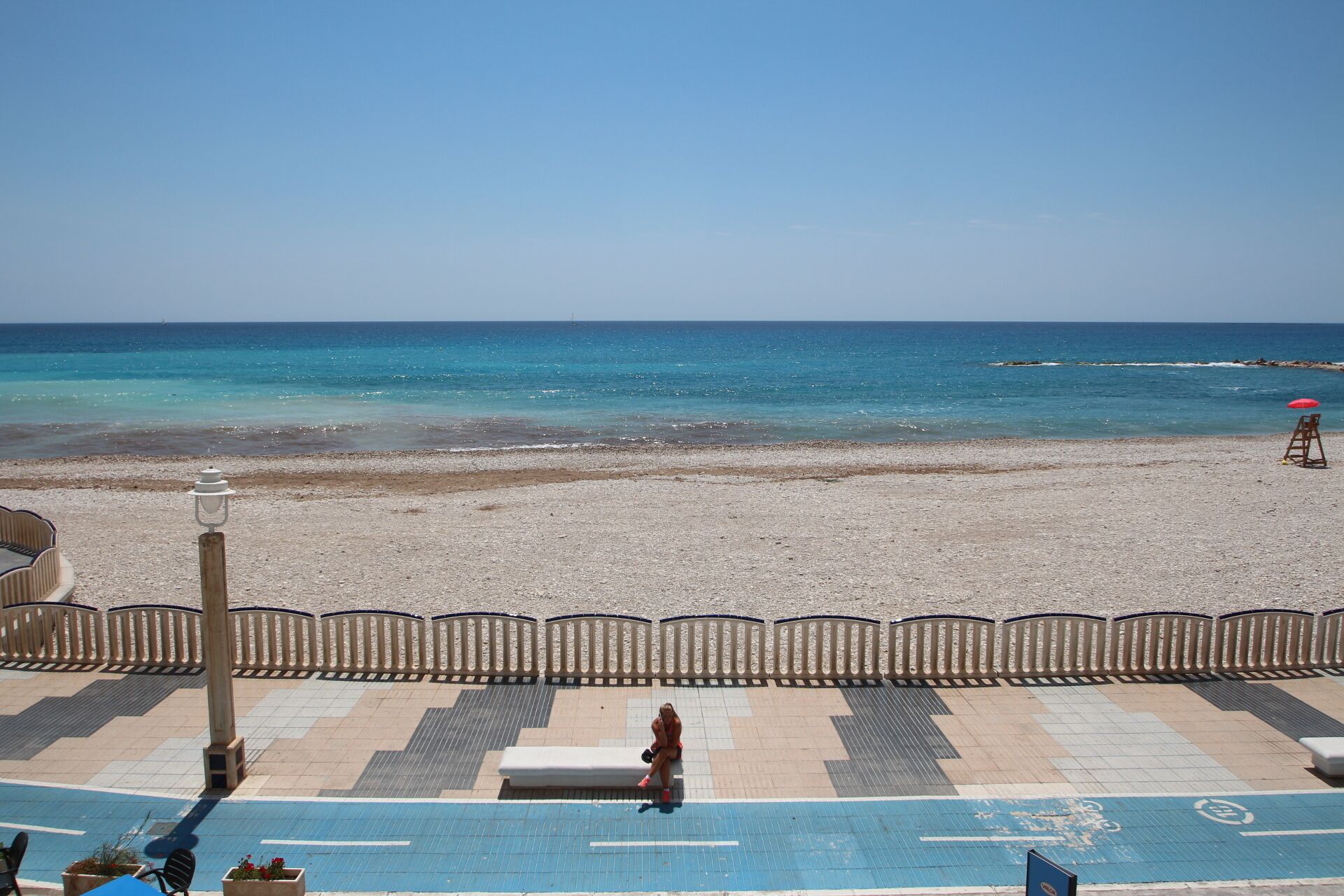 Une plage avec une chaise de maître-nageur et un parasol rouge, une promenade dallée et un banc.