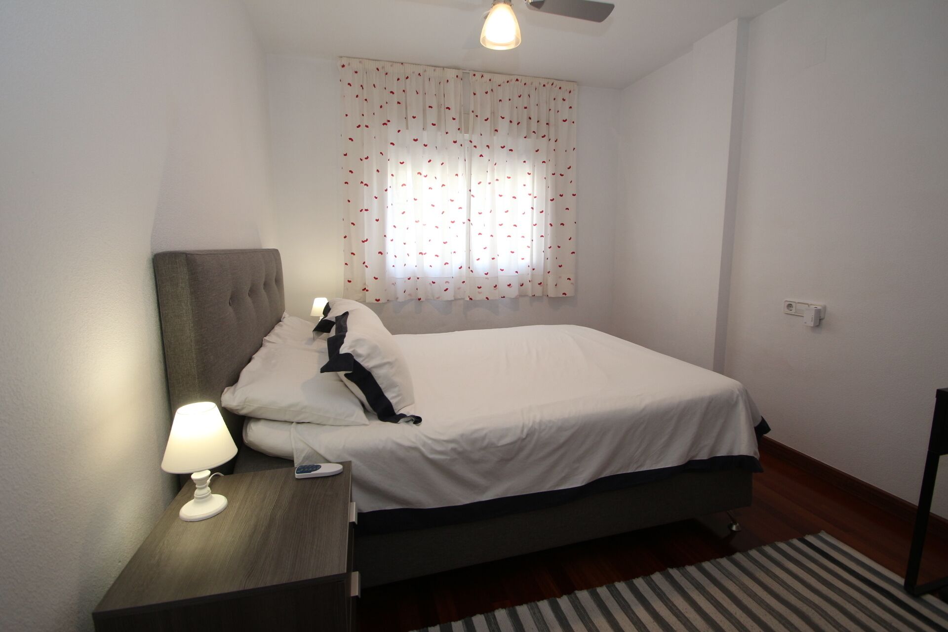 A neatly arranged bedroom with a bed, bedside table, lamp, and a window with heart-patterned curtains.