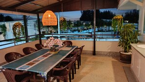 A dining area with a table set for four, surrounded by chairs, a decorative centerpiece, and a view of the street through large windows. - Wonderful Villa with Private Pool, Best Location! Walk to beach, restaurants etc (Khao Lak)