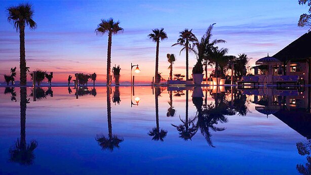 A serene pool area with palm trees and a sunset. - Sun-Drenched, Sea-View Apartment with Giant Patio, Private Garden and Garage (Fuengirola)
