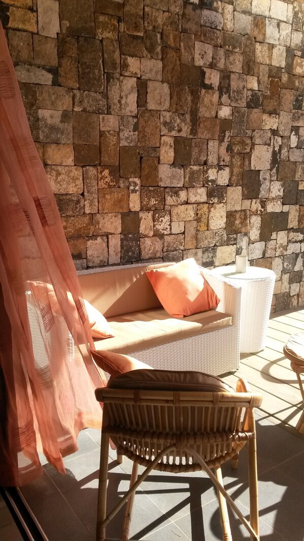 Una silla de mimbre y un banco acolchado con una cesta tejida a su lado, frente a una pared de piedra.