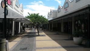 A pedestrian walkway with storefronts on either side, a tree in the center, and a clear sky.