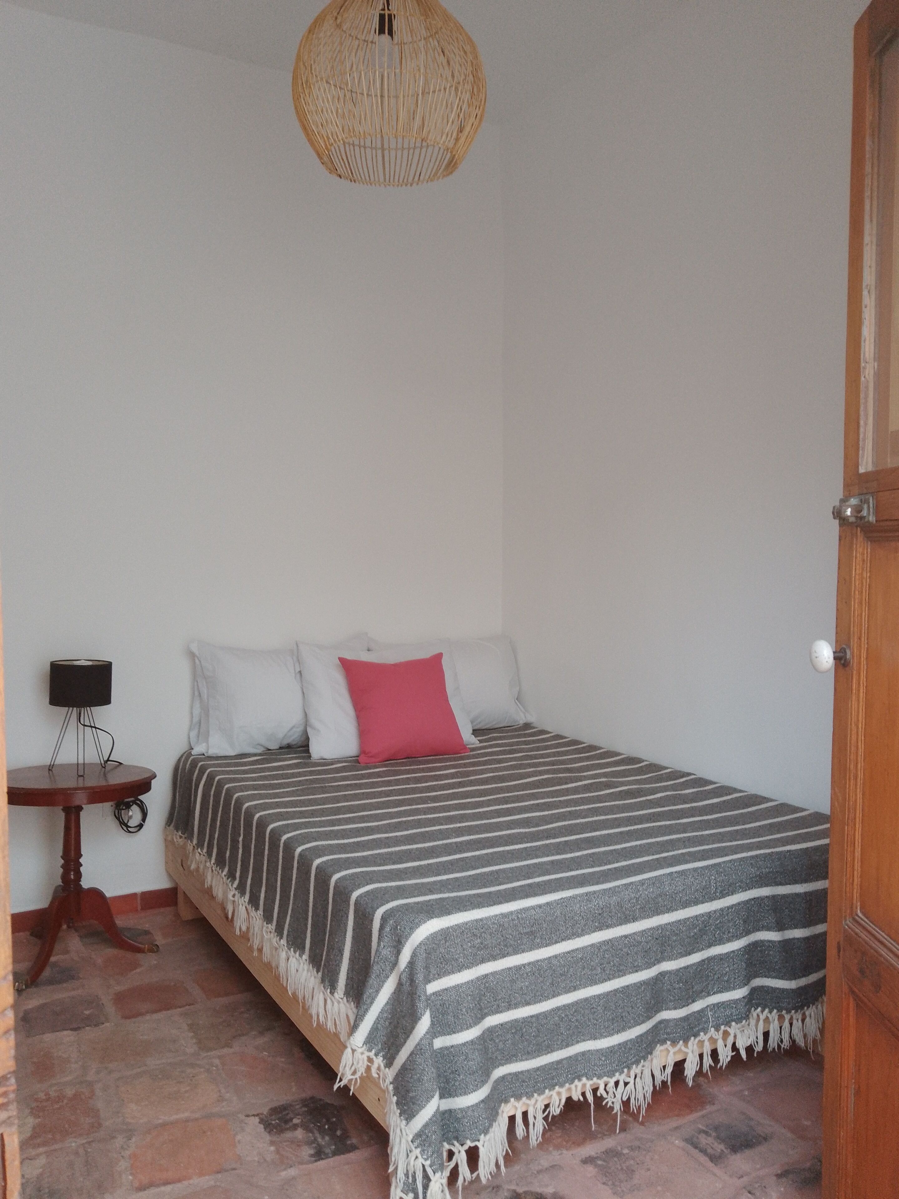 A bedroom with a bed, a bedside table, a lamp, and a wicker ceiling light.