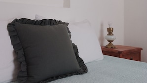 En seng med en mørkegrå pude og en hvid pude, et let blåt, struktureret tæppe, et trænatbord med en messinglampe, og en hvid væg med et simpelt buet design.
