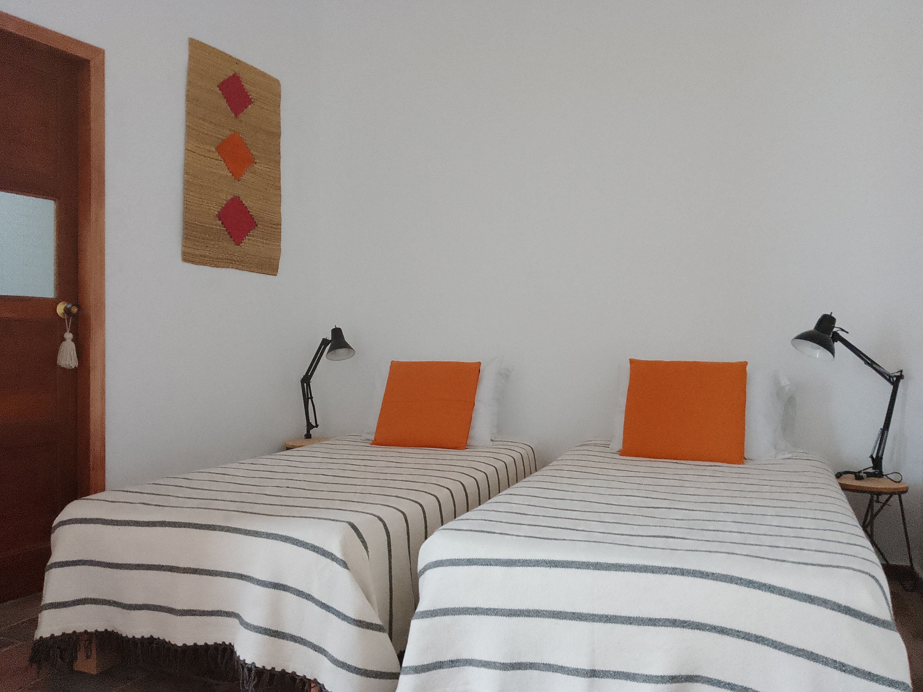 Een kamer met twee enkele bedden, elk voorzien van gestreept beddengoed en een oranje kussen. Twee nachtkastjes met lampen, en een wanddecoratie met een geometrisch patroon.