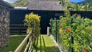 A rustic building with a stone wall and a wooden fence, surrounded by lush greenery and a mountain backdrop.