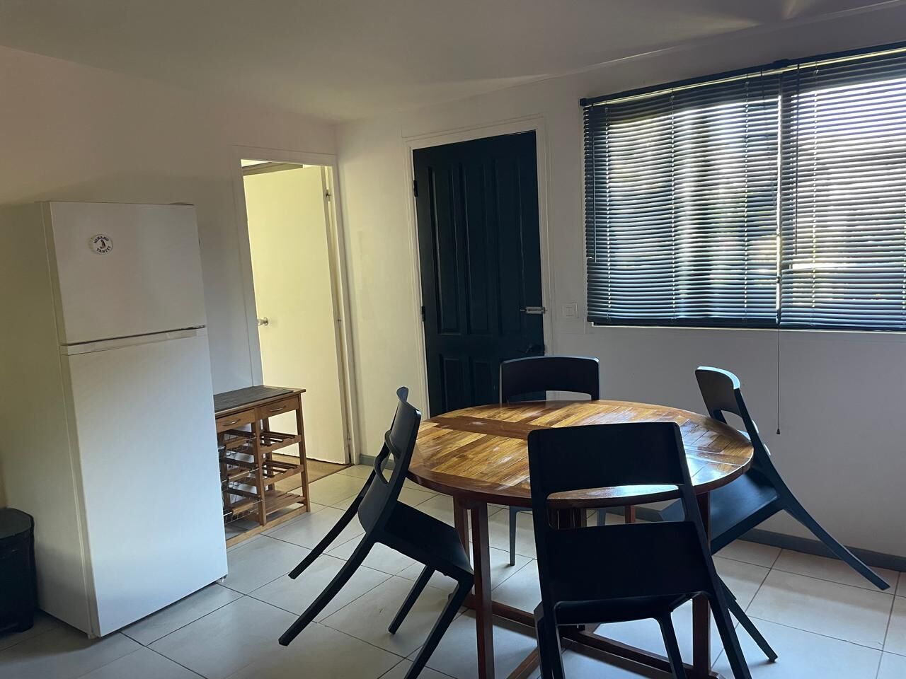 Un ambiente abitativo compatto con un tavolo da pranzo rotondo in legno, circondato da sedie nere, un frigorifero bianco e un piccolo tavolino in legno.