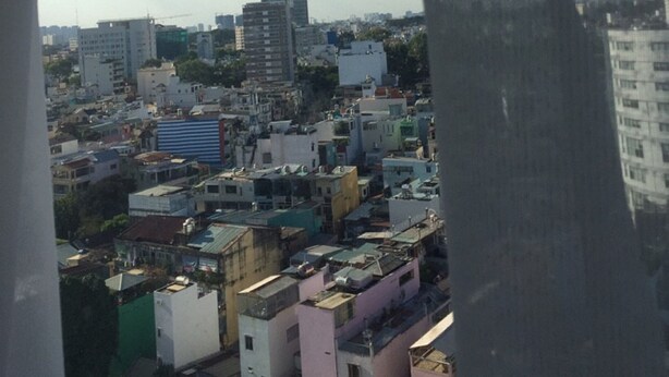 Une vue sur un paysage urbain, avec différents bâtiments, dont des immeubles de grande et de petite hauteur, observée à travers une fenêtre ornée d’un rideau léger et transparent.