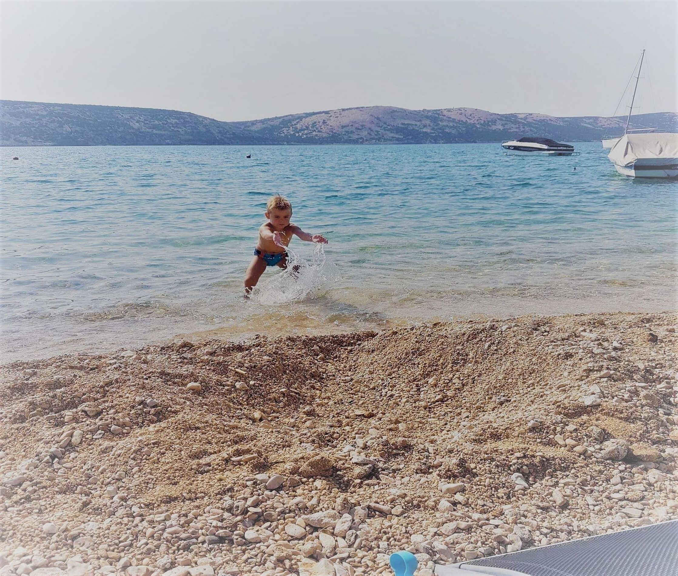 A child playing in shallow water near a pebble beach with boats in the background.