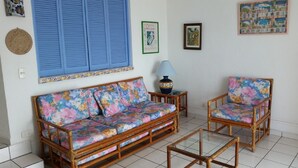 Una sala de estar con un sofá y dos sillones, una lámpara y adornos de pared, incluyendo un cuadro enmarcado y un cartel.
