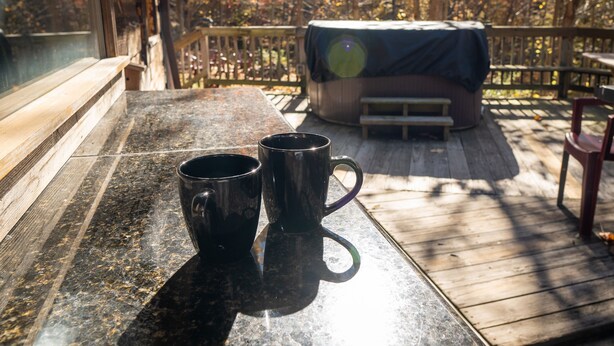 Eine Holzterrasse mit zwei schwarzen Tassen auf einem Tisch, einem Grill mit Deckel und einer Bank.