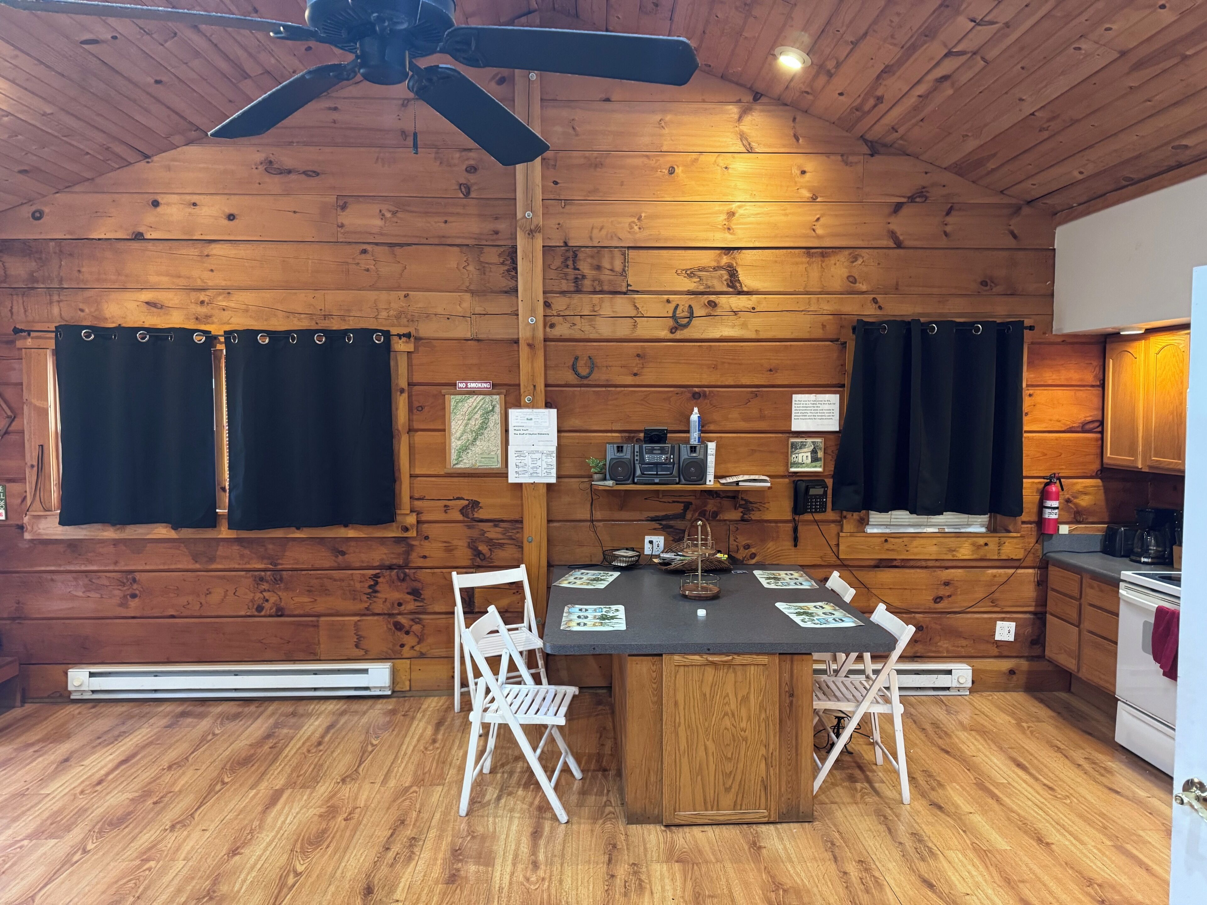 A wooden cabin interior with a dining table, chairs, a ceiling fan, and a kitchen area.