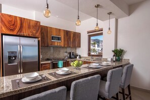 A modern kitchen with a stainless steel refrigerator, a microwave, and a stove. There are bar stools and a large island with a bowl of fruit.