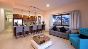 A modern kitchen with a dining area, a living room with a sofa and a coffee table, and a large window with a city view.