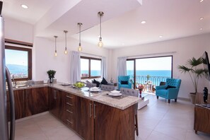 Une cuisine moderne dotĂ©e dâun Ăźlot central, dâun coin repas avec des chaises bleues et de grandes fenĂȘtres offrant une vue sur la mer.