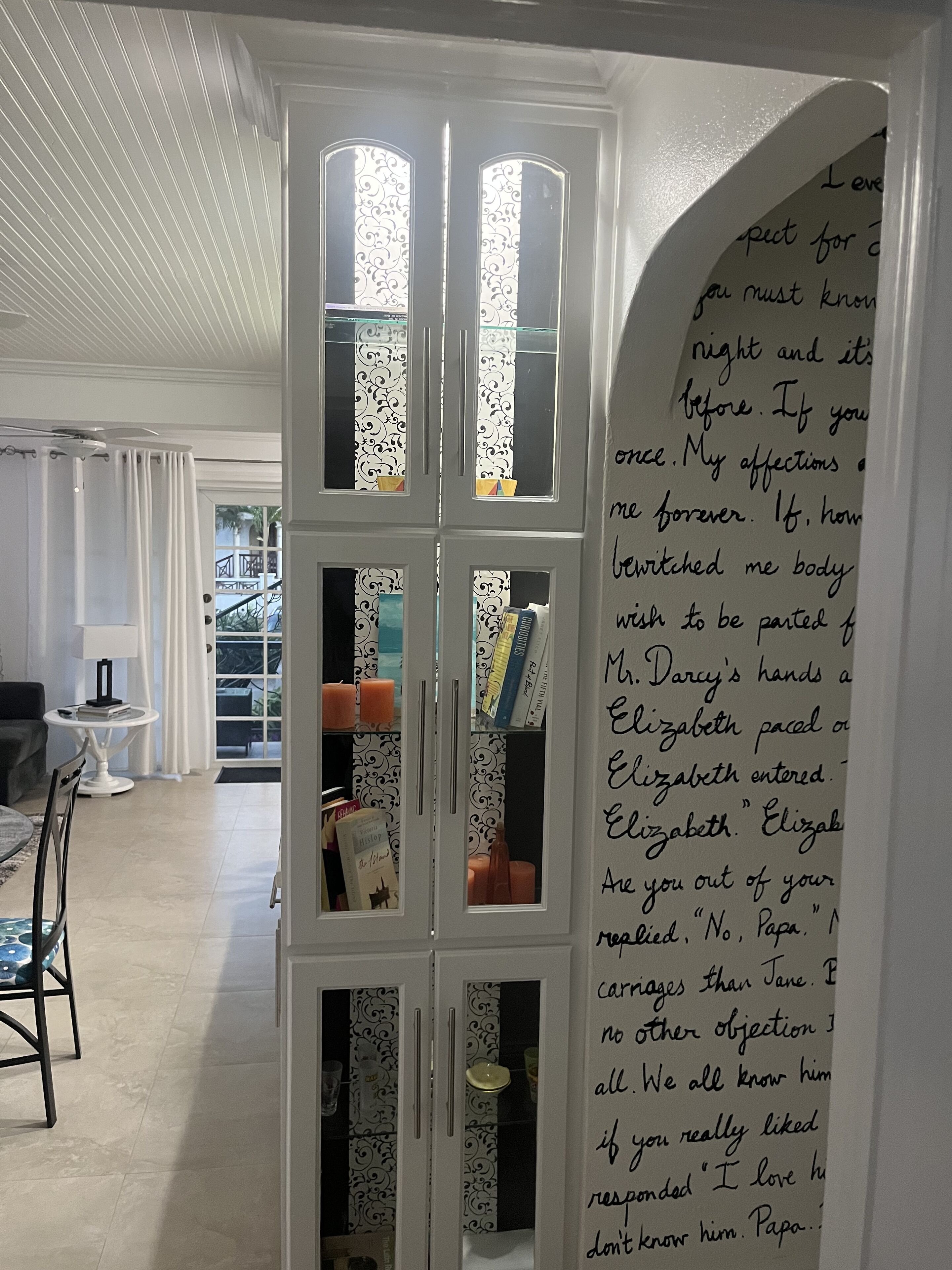 A room with a white cabinet containing books and candles, a dining table with chairs, and a view of a balcony with outdoor furniture.