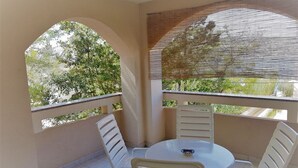 Een balkon met een ronde tafel en witte stoelen, uitzicht op bomen en een bamboe zonnescherm.