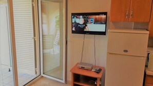 Een kamer met een schuifdeur naar buiten, een aan de muur bevestigde televisie, een klein houten tafeltje met een dvd-speler en een witte koelkast.