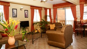Una sala de estar con sofá, sillón, mesa de centro y televisor. Hay cortinas, un ventilador de techo y una zona de comedor al fondo.