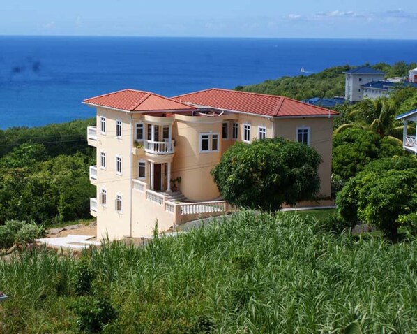 A two-story building with a red roof, surrounded by greenery and overlooking a body of water.