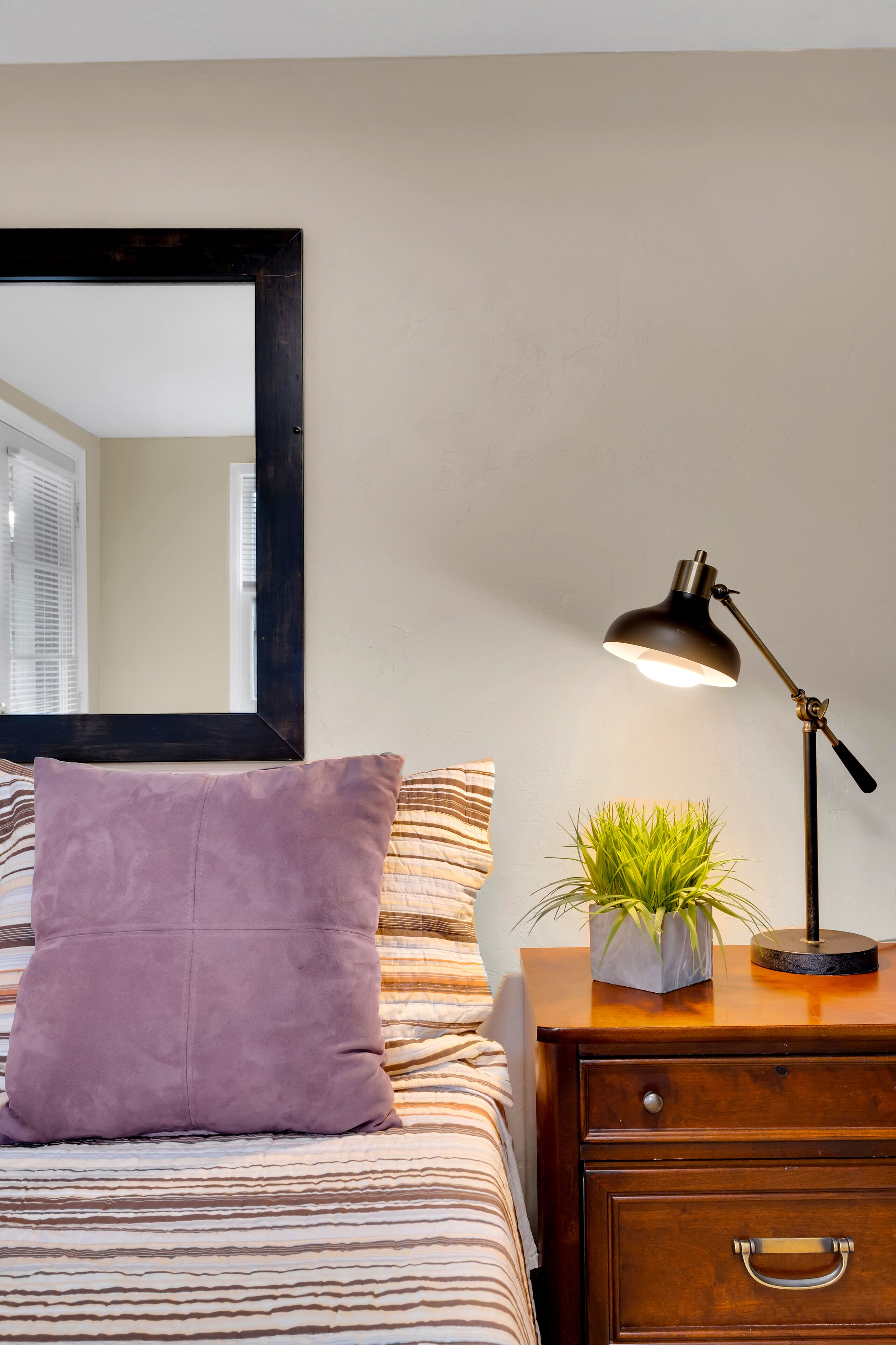 A neatly made bed with striped bedding, a purple pillow, a bedside table with a lamp, and a small potted plant.