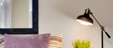 A neatly made bed with striped bedding, a purple pillow, a bedside table with a lamp, and a small potted plant.