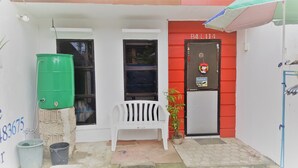 Entrada de un edificio con una puerta roja identificada con la etiqueta "B4 L114", un banco blanco, un contenedor verde y una planta en maceta.