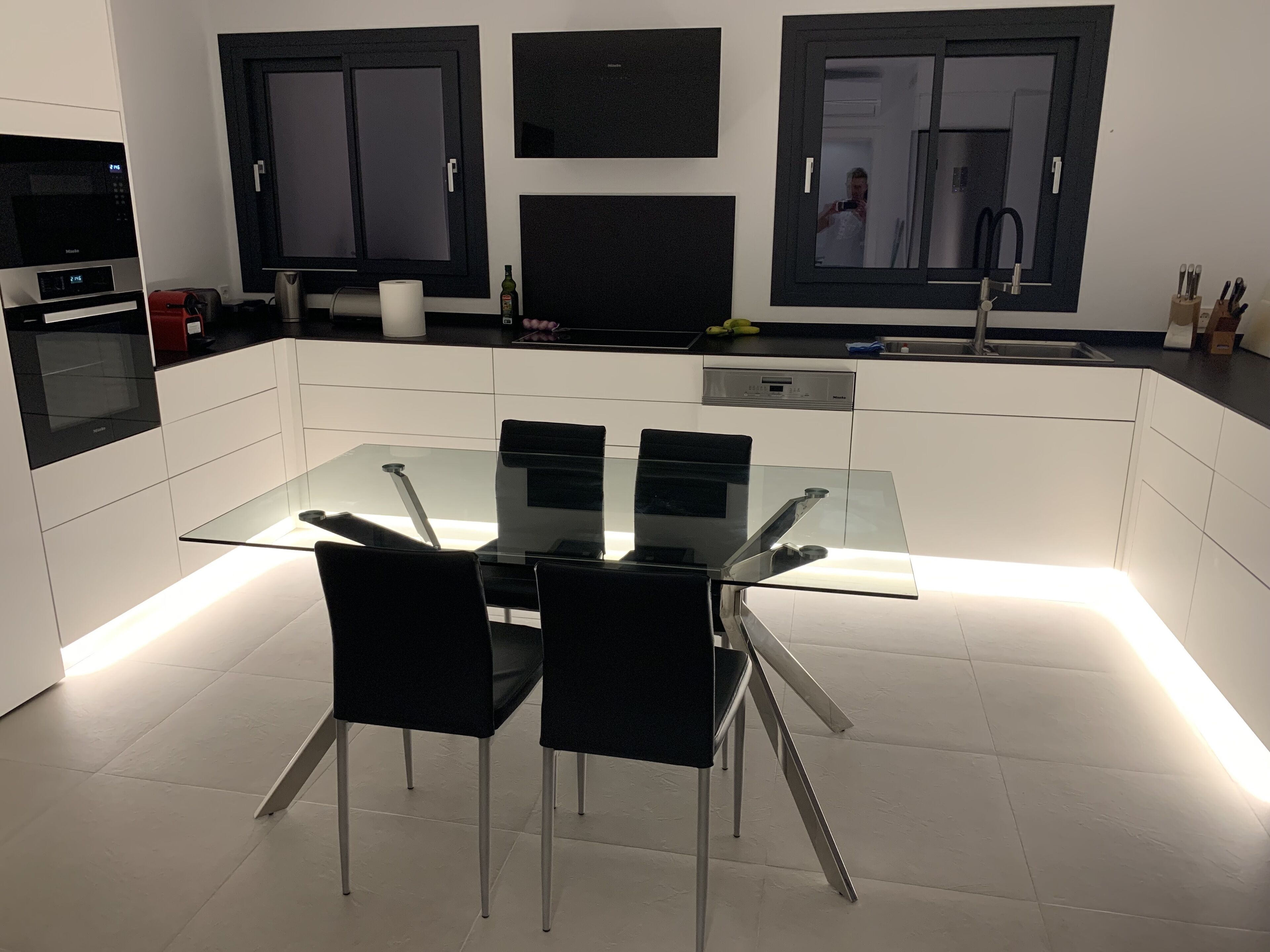 A modern kitchen with a glass dining table, black chairs, and white cabinetry.