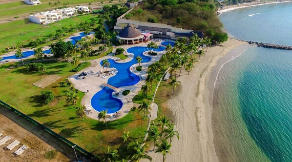 An aerial view of a resort with multiple swimming pools, a beach, and a large building with a thatched roof. - Beach Break Villa inside B Nayar's Beach Club & gated Community. (Cruz de Huanacaxtle / Bucerias)