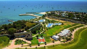 Vista aérea de un complejo turístico frente al mar, con playa, zona de piscina y canchas deportivas.