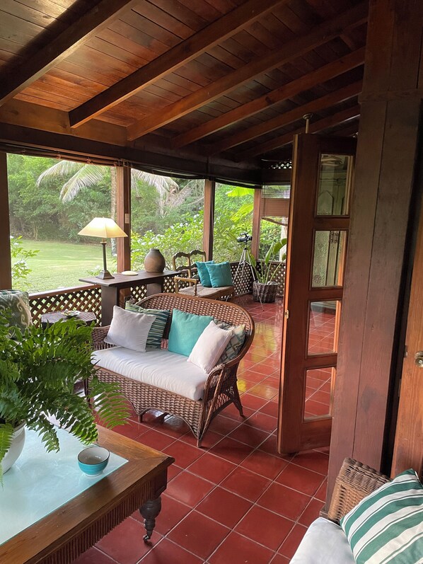 Relax on the veranda with a serene garden view, featuring a wicker sofa and a table with a lamp.