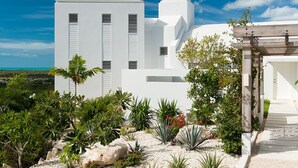 Un edificio moderno blanco con techo plano, rodeado de plantas tropicales y un cielo azul despejado.