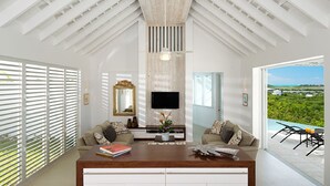 Un salón amplio con techo alto, un televisor de pantalla plana en la pared y vista a un paisaje exuberante a través de grandes ventanales.