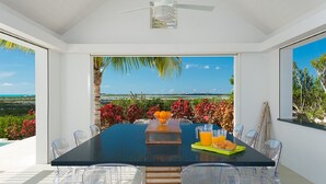 Un comedor con una mesa, sillas, ventilador de techo y vistas al océano y la vegetación.