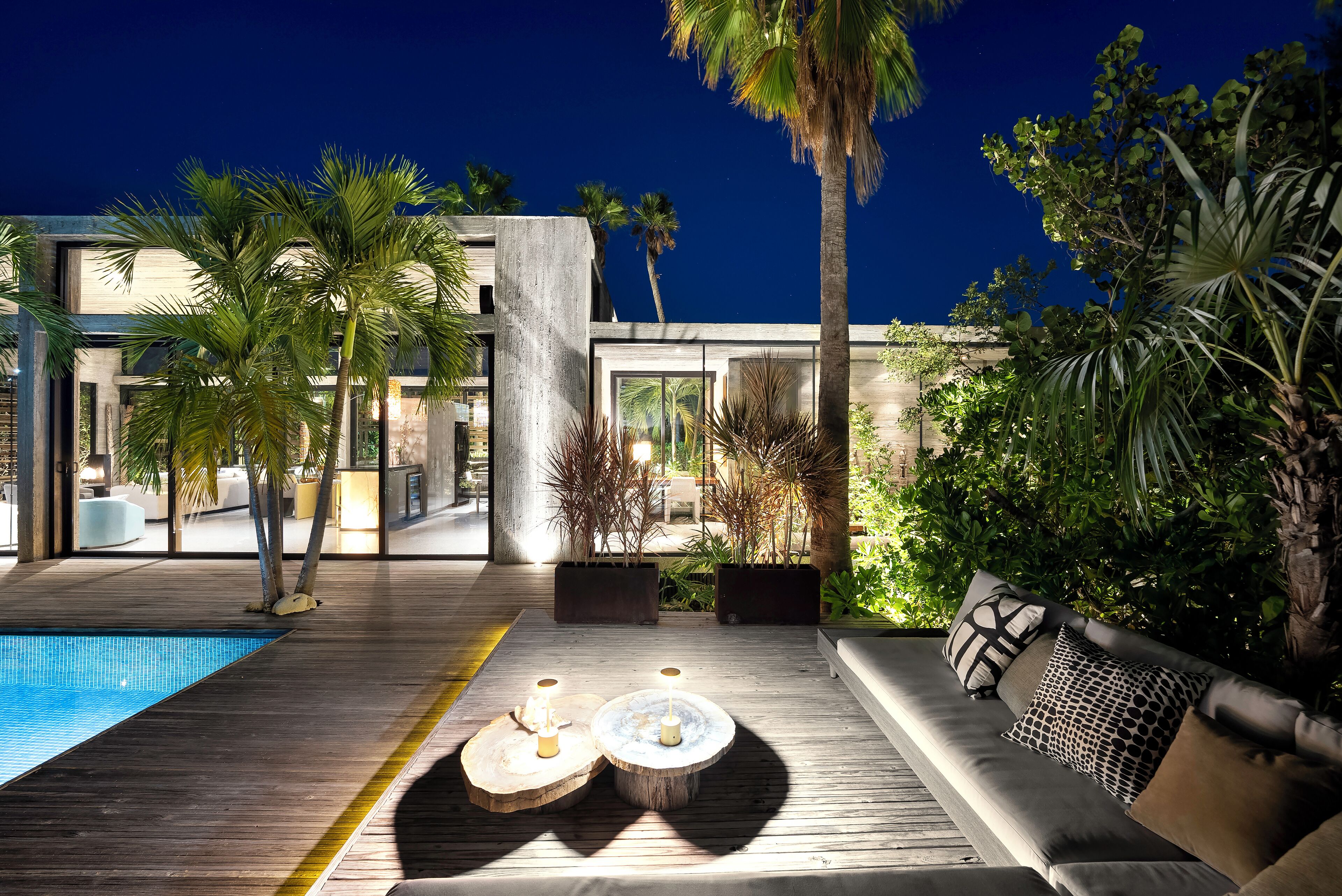Un espace piscine extérieur moderne, doté d’une terrasse en bois, d’un salon avec un canapé et une table basse, et de palmiers.